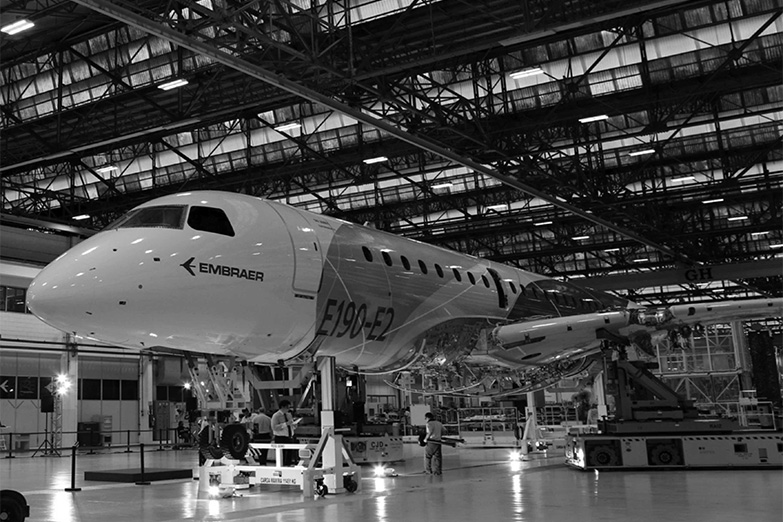 Foto em preto e branco mostrando uma aeronave grande no hangár com o logo da Embraer